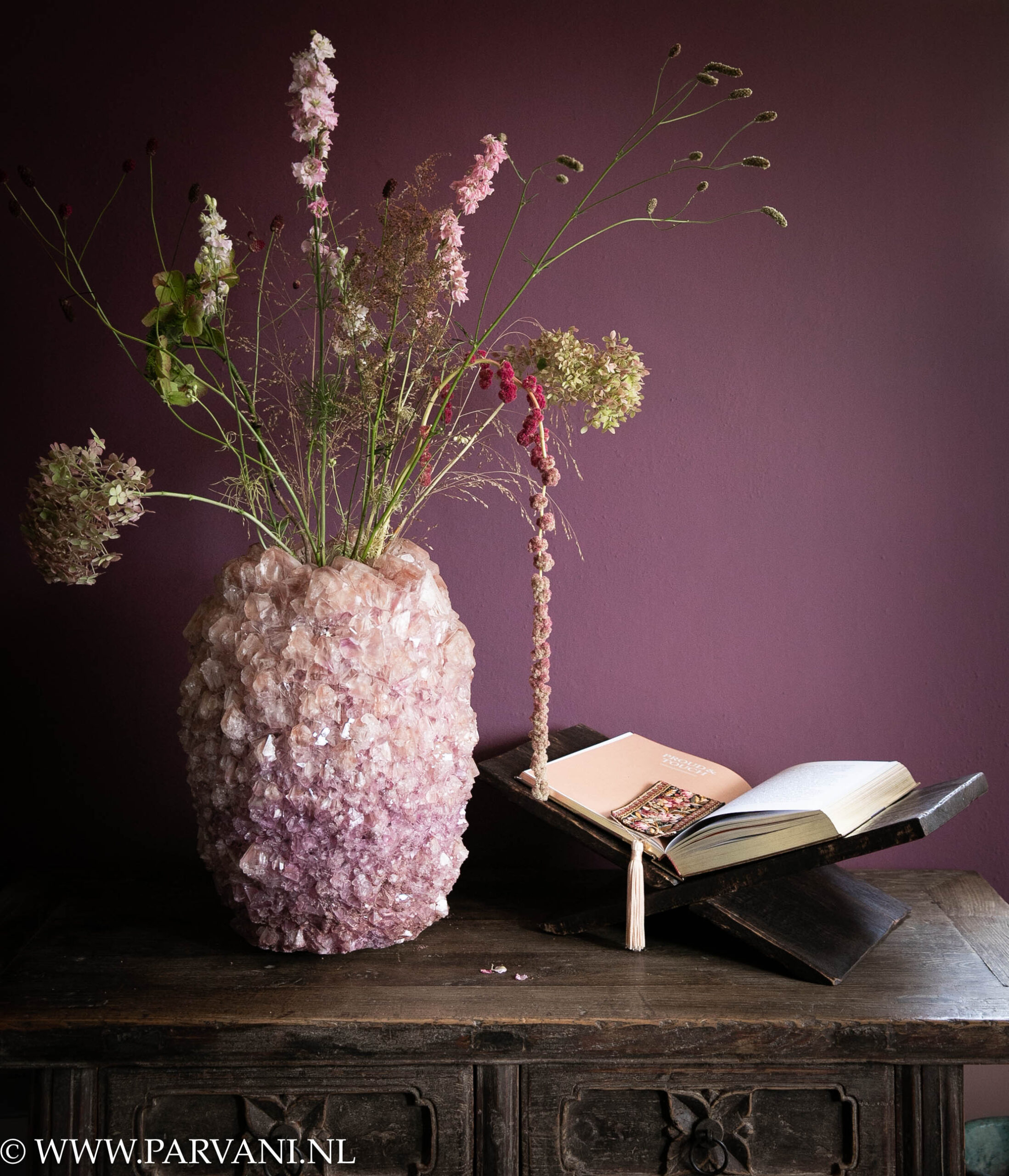 Grote roze paarse mineraal kristallen vaas met luchtig boeket en donkerbruine houten boekenstandaard