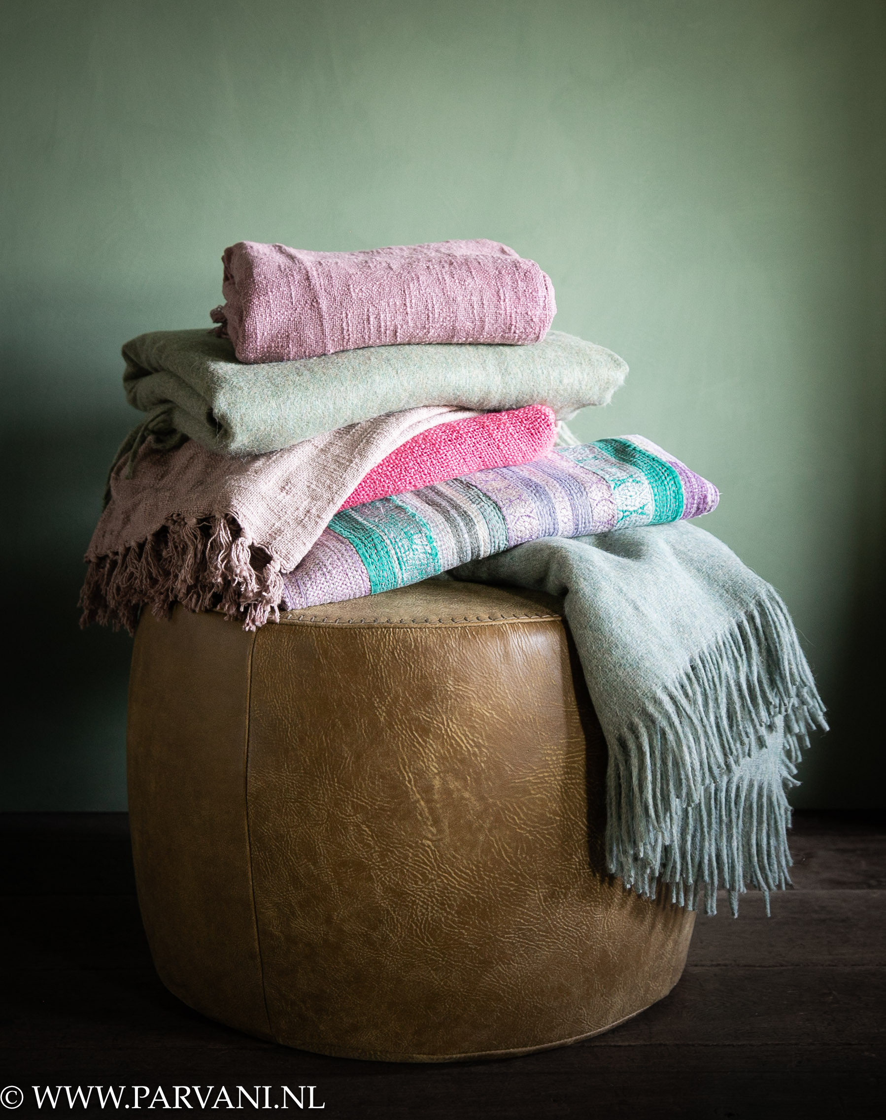 Stapel kleurrijke plaids in frisse groen, roze en lila paarse kleuren op stoere ronde poef van leer